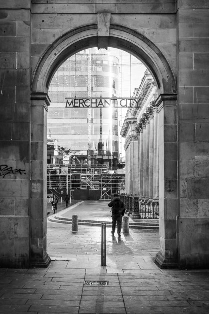 A black and white portrait orientation image looking through a stone archway towards Royal Exchang Square in Glasgow, with a glass building in the background and a sign saying Merchant City near the top of the archway. A man is walking away from the camera on the other side of the archway and his shadow is being cast backwards towards the viewer.