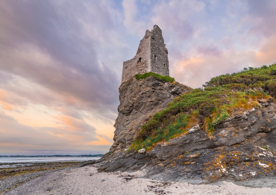 An image looking up at a castle ruin on a rocky cliff from the beach, with a dramatic cloudy sky full of oranges, yellows, pinks, and blues.