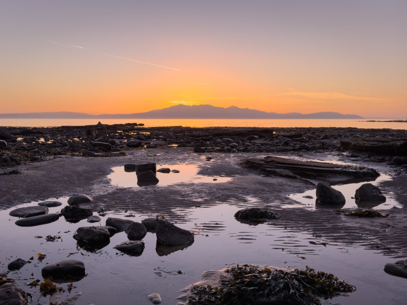 a coastal scene with a beach and rocks with pools of water reflecting the vivid blues and oranges of the sky, with an island in the background across and expanse of water, the sun setting just behind the mountain range that dominates the silhouette of the island.